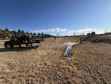  Biolein crew member flushing a concrete drainage channel with a vacuum truck hose on an open grass field 