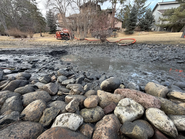  Close-up of dredged pond sediment along a rocky shoreline with vacuum trailer staged in the background 